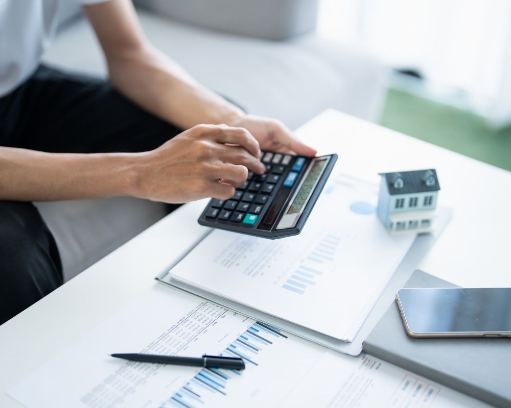 Persona usando calculadora junto a documentos financieros y maqueta de casa